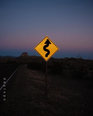 A yellow traffic sign warns of a winding road ahead at dusk, with a dark asphalt road and hilly landscape visible.