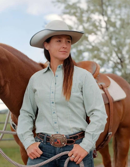 A woman in western attire stands in front of a horse wearing a saddle, outdoors on a sunny day. The woman wears a cowboy hat.