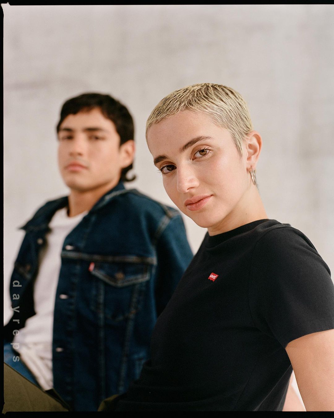Close-up of a short blonde-haired woman in a black t-shirt with a man in a denim jacket blurred in background.