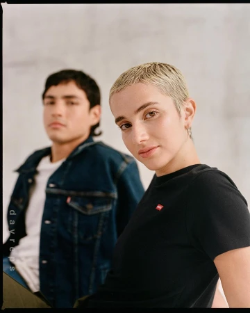 Close-up of a short blonde-haired woman in a black t-shirt with a man in a denim jacket blurred in background.