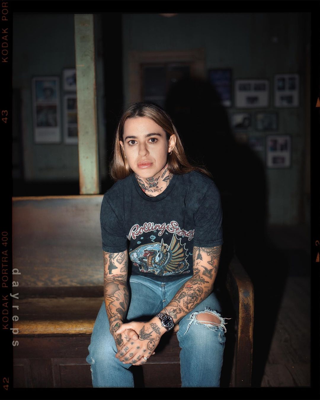 Portrait of a tattooed young adult woman with blonde hair, sitting on a wooden bench indoors, wearing a Rolling Stones t-shirt and jeans.