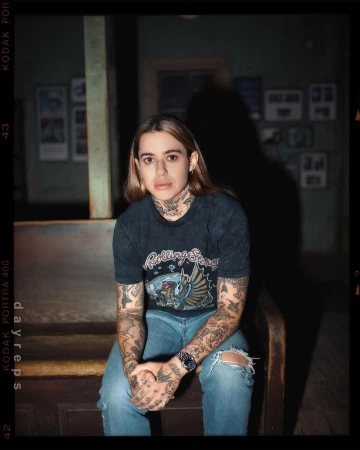 Portrait of a tattooed young adult woman with blonde hair, sitting on a wooden bench indoors, wearing a Rolling Stones t-shirt and jeans.
