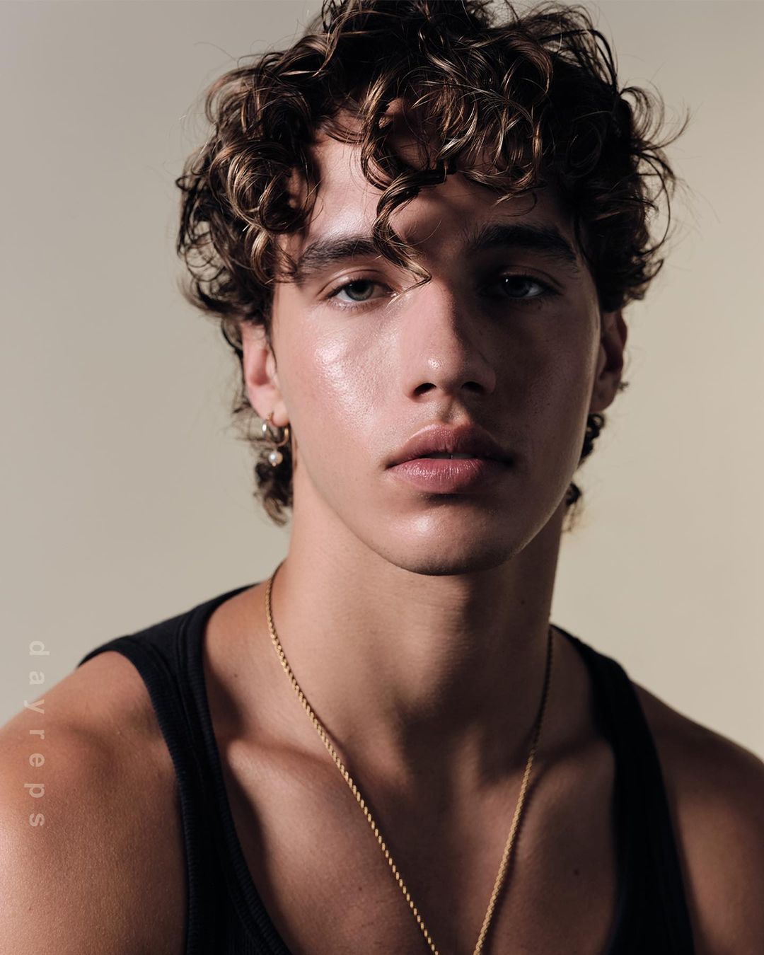 Portrait of a young adult male with curly brown hair wearing a black tank top and a gold chain necklace against a neutral background.