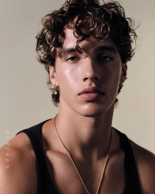 Portrait of a young adult male with curly brown hair wearing a black tank top and a gold chain necklace against a neutral background.