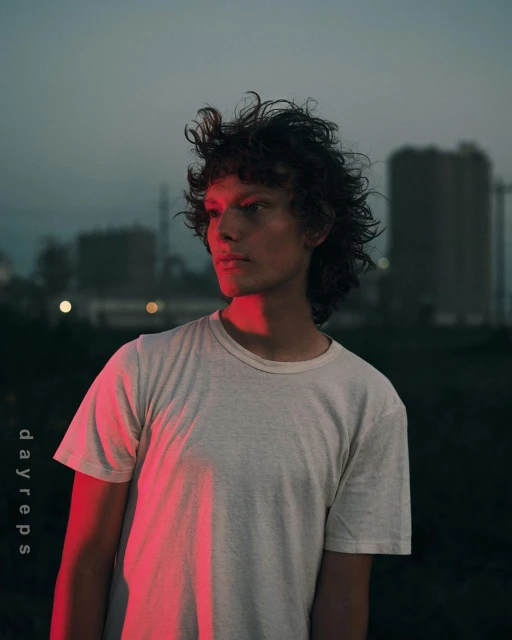 Portrait of a young man with curly hair, wearing a t-shirt, set against an industrial backdrop under a twilight sky with soft, red lighting.
