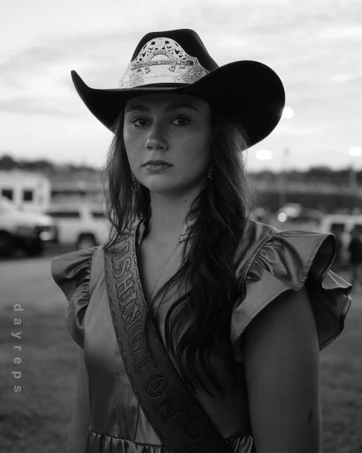 Portrait of a young woman wearing a cowboy hat and sash, captured in black and white at an outdoor event with cars in the background.