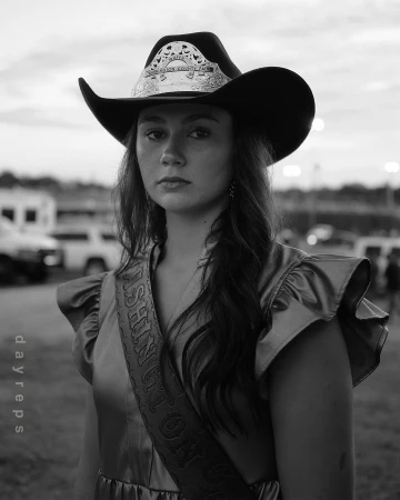 Portrait of a young woman wearing a cowboy hat and sash, captured in black and white at an outdoor event with cars in the background.