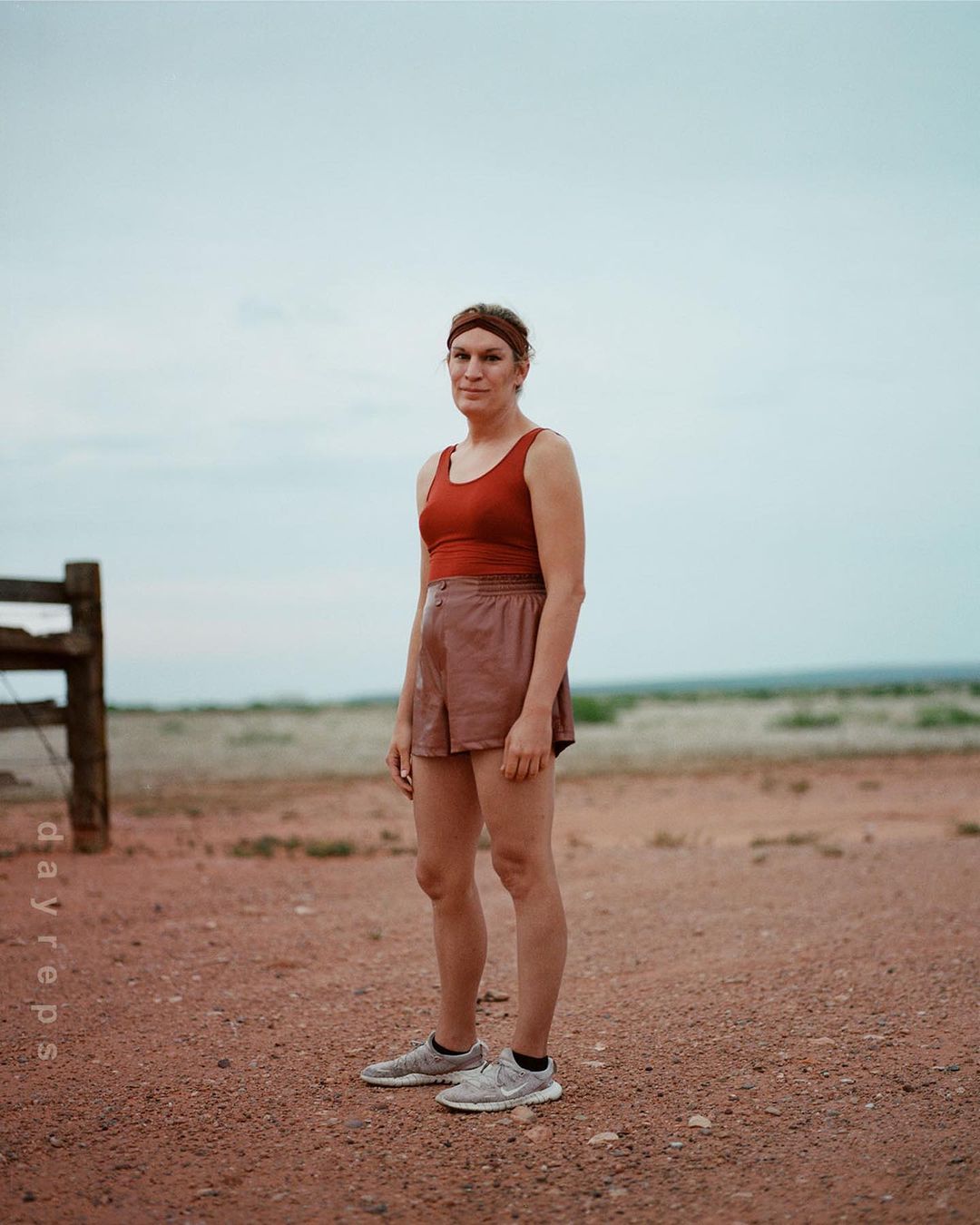 Adult female in sportswear stands outdoors against a flat horizon on a dirt surface, captured in a full shot with natural lighting.