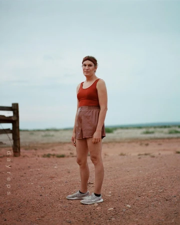 Adult female in sportswear stands outdoors against a flat horizon on a dirt surface, captured in a full shot with natural lighting.