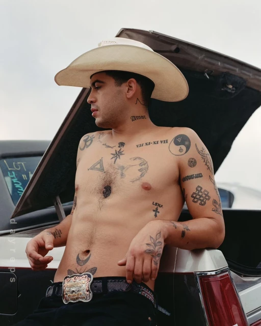 Tattooed man in cowboy hat leans against classic car trunk, bare-chested with ornate belt buckle, outside in soft light.