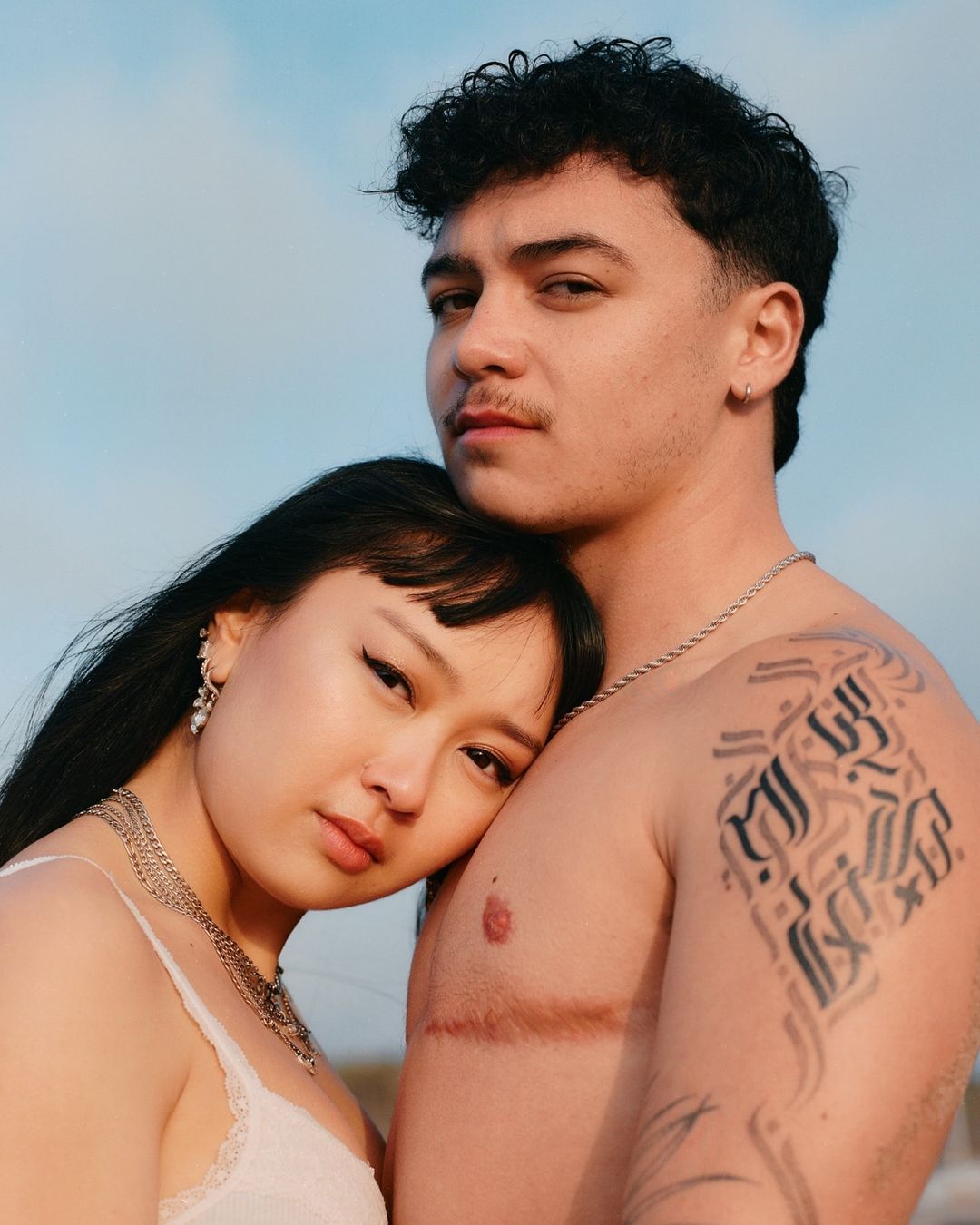 Intimate portrait of a young adult couple embracing in natural light, with the sky as a backdrop, highlighting their connection and details.
