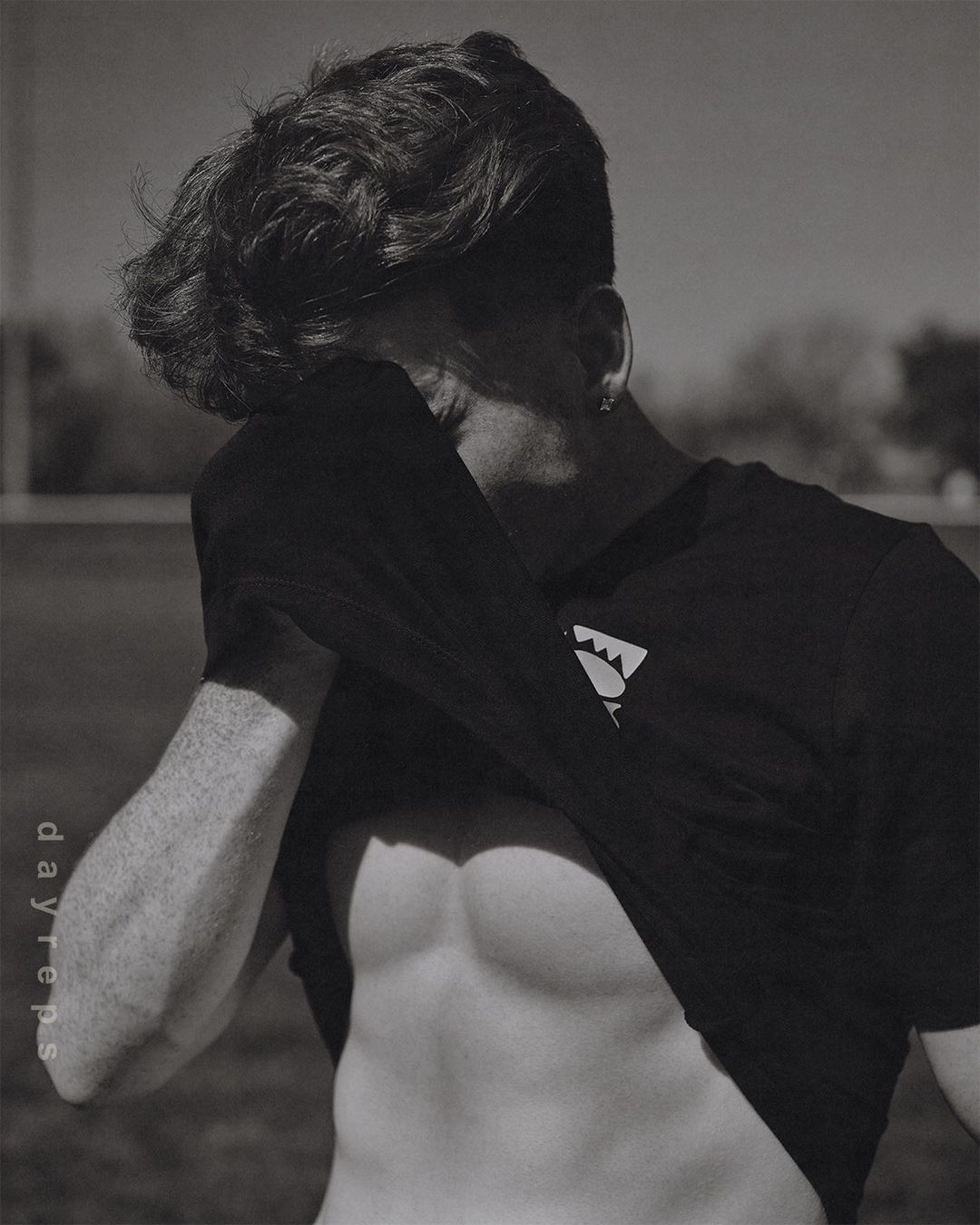 Black and white portrait of muscular adult male with exposed abs, wearing t-shirt in outdoor setting with blurred background.