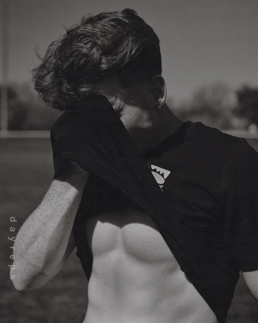 Black and white portrait of muscular adult male with exposed abs, wearing t-shirt in outdoor setting with blurred background.
