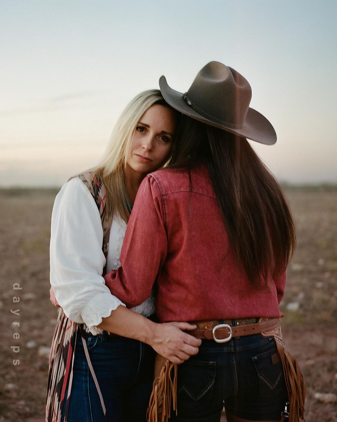 Two adult women embrace outdoors in a field at dusk. One woman wears a cowboy hat, short denim shorts, a red shirt, and a leather chaps.