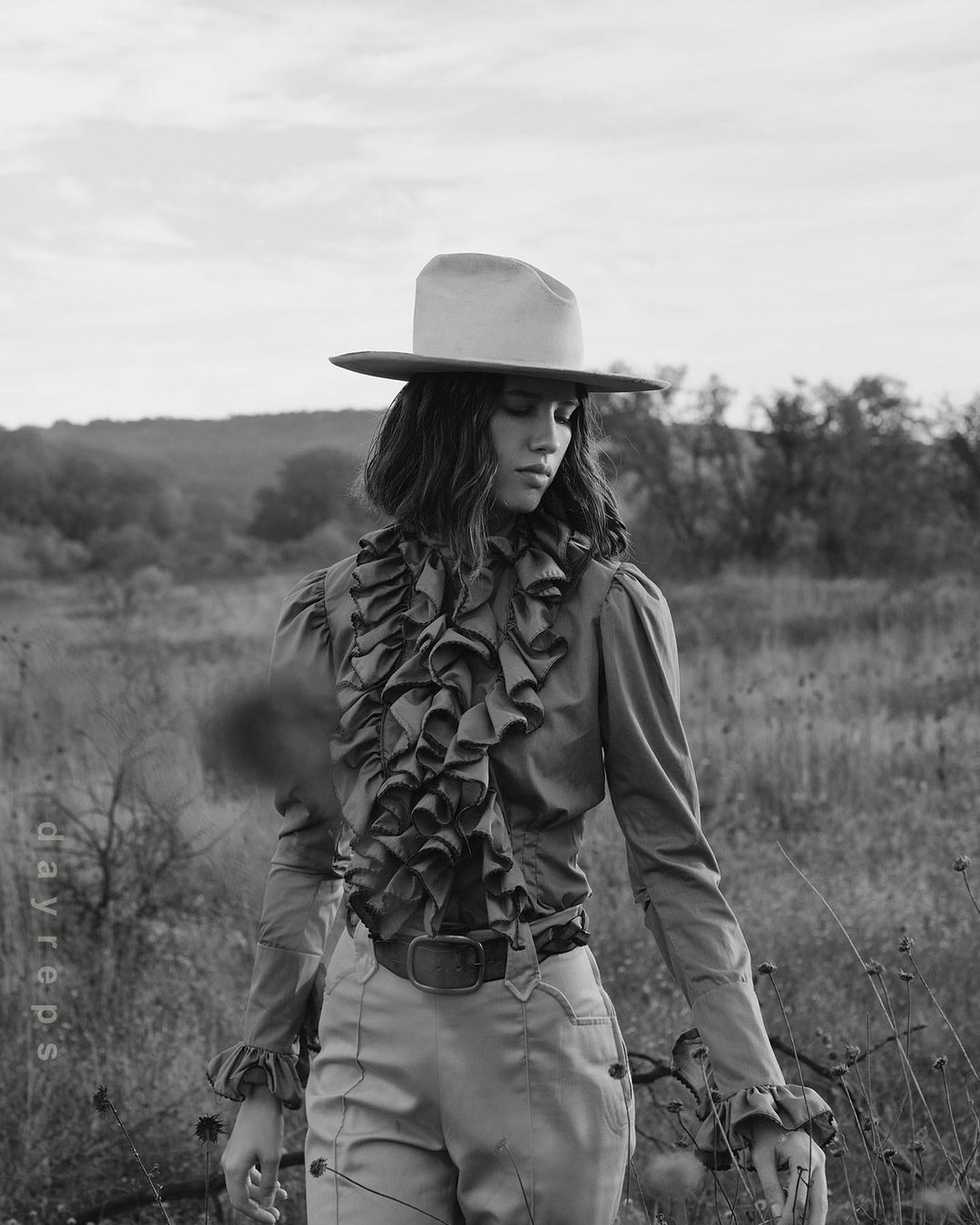 Monochrome portrait of a woman in a hat and ruffled shirt standing in a field with a blurred landscape background, captured in a vintage editorial style.