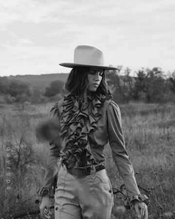 Monochrome portrait of a woman in a hat and ruffled shirt standing in a field with a blurred landscape background, captured in a vintage editorial style.