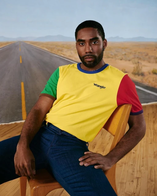 Man in a yellow and red Wrangler t-shirt sits on a chair against a desert highway backdrop, showcasing fashion apparel.