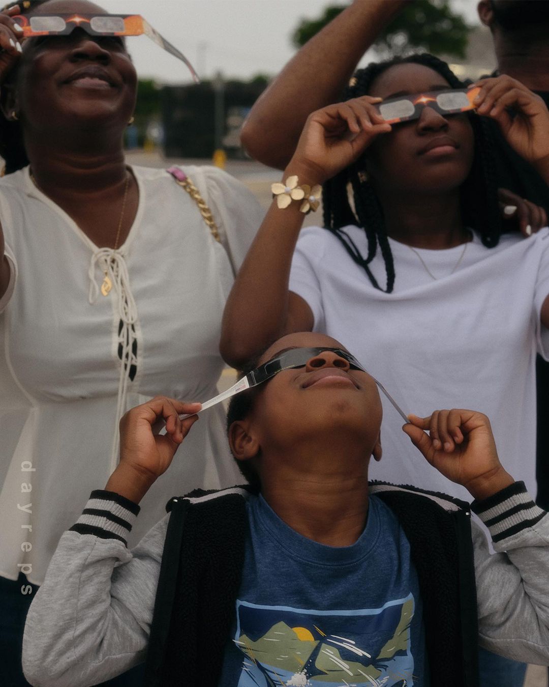 Three people wear solar eclipse glasses and look up at the sky. An adult woman and two children are partially visible in the outdoor daytime shot.