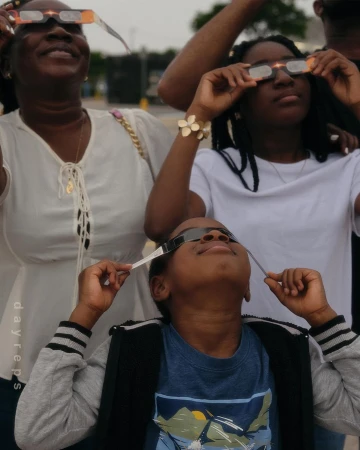 Three people wear solar eclipse glasses and look up at the sky. An adult woman and two children are partially visible in the outdoor daytime shot.