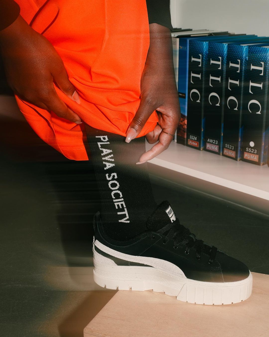 Close-up of a model wearing black Puma platform sneakers, black leggings with 'Playa Society' text, and orange shorts indoors.