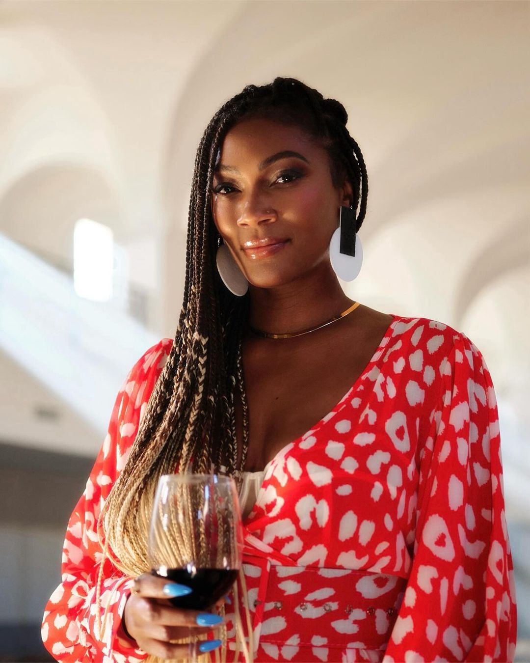 Adult woman in a patterned red dress holds a wine glass indoors, posing with arched architecture in the background.