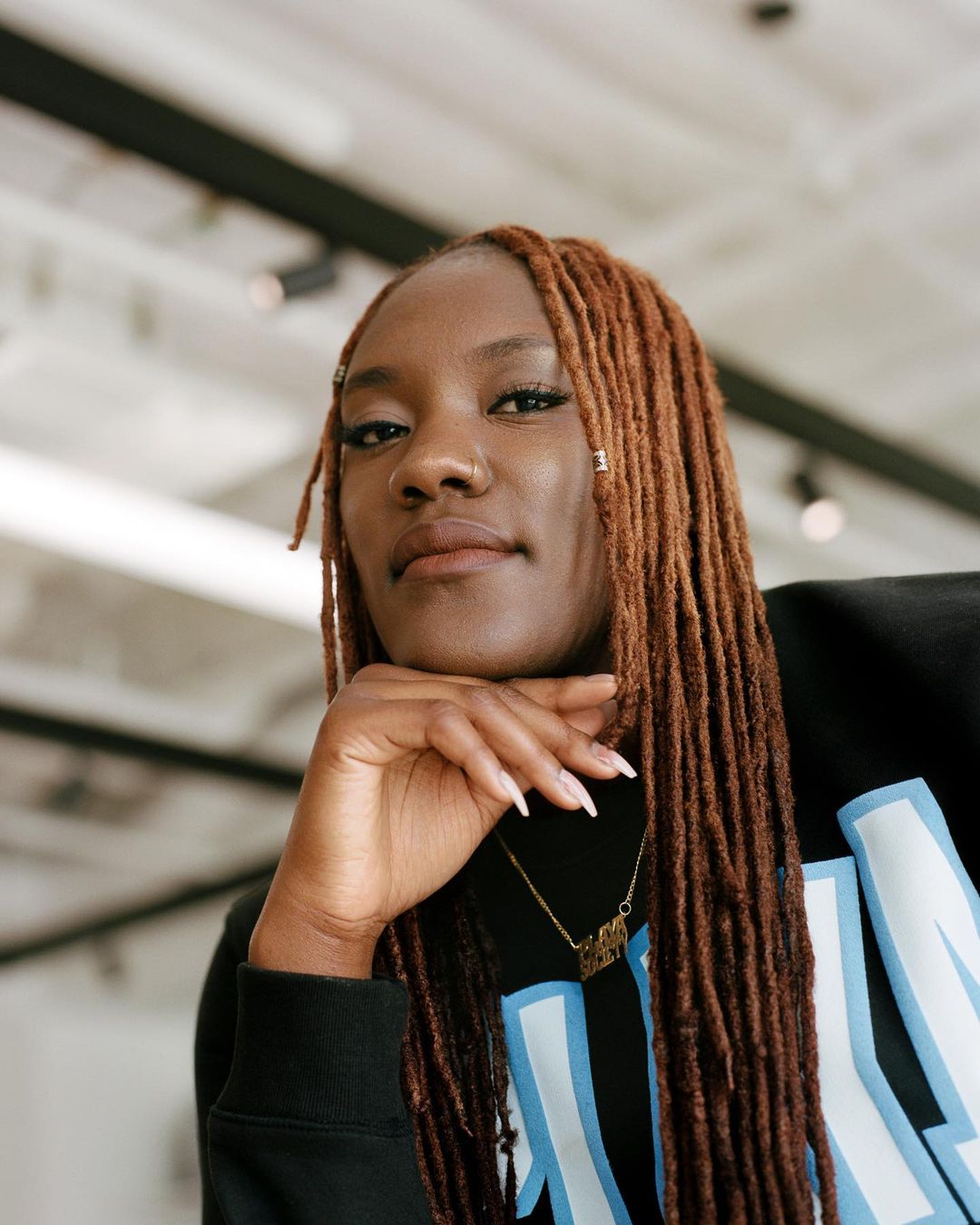 Portrait of a Black woman with locs, wearing a black sweatshirt, posing indoors with her chin on her hand, looking at the camera.