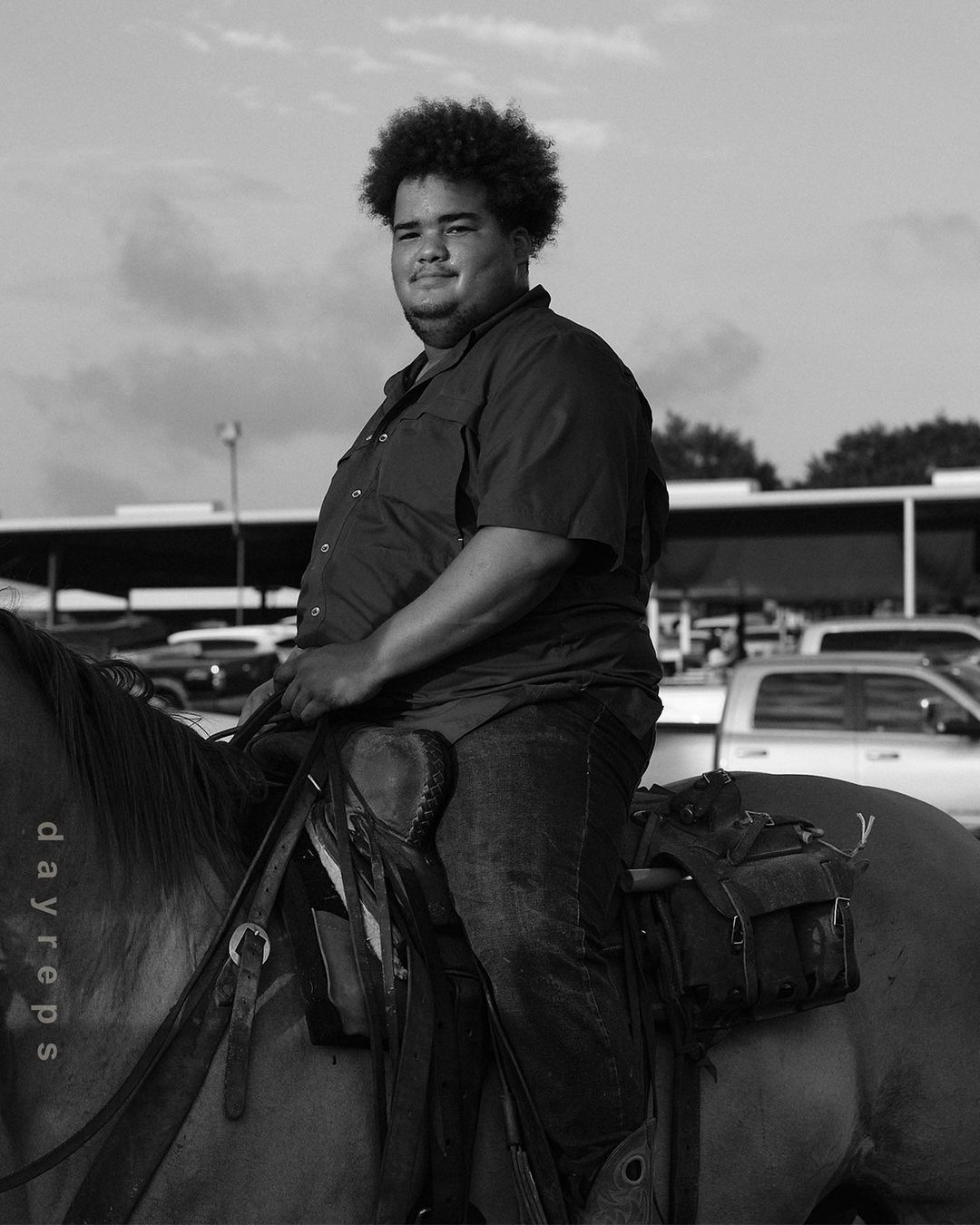 A dark-haired adult male smiles while seated on a horse outdoors. The monochrome image features a bright sky and parked vehicles.
