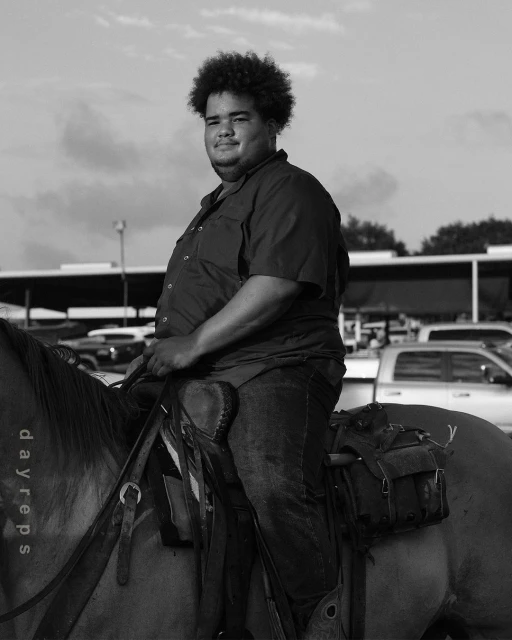 A dark-haired adult male smiles while seated on a horse outdoors. The monochrome image features a bright sky and parked vehicles.