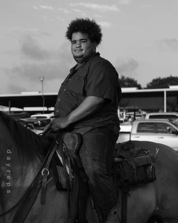 A dark-haired adult male smiles while seated on a horse outdoors. The monochrome image features a bright sky and parked vehicles.