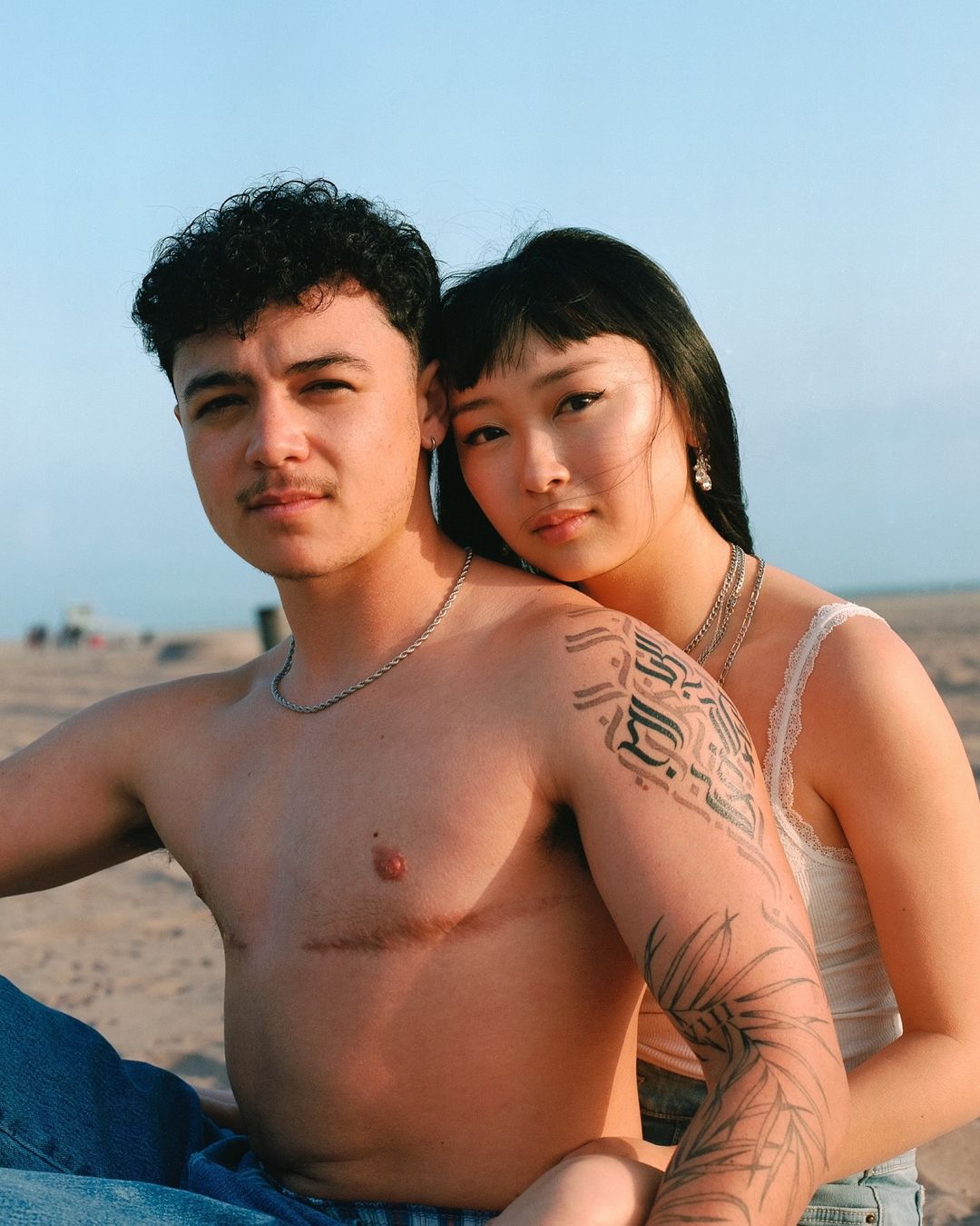 A close-up of a couple sitting on a beach, with the man shirtless and the woman leaning on him, with blue sky in the background.
