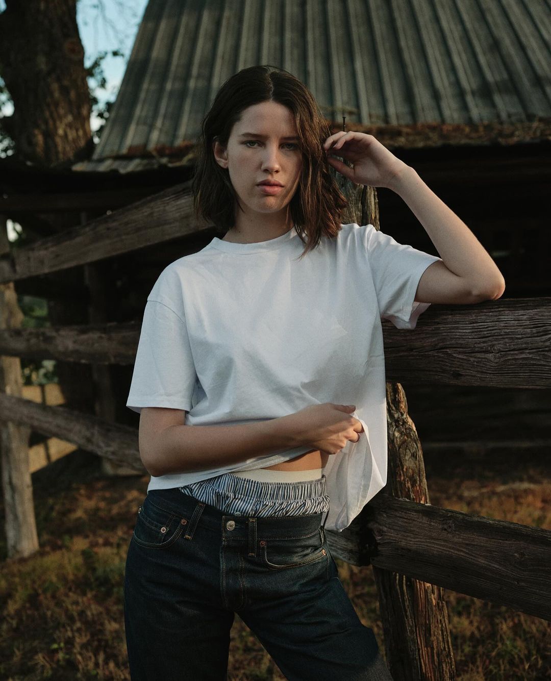 Young woman with short brown hair poses against a rustic wooden fence in a relaxed outdoor fashion portrait on a sunny day.