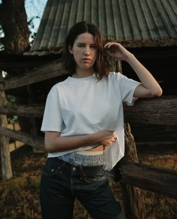 Young woman with short brown hair poses against a rustic wooden fence in a relaxed outdoor fashion portrait on a sunny day.