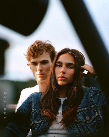 Close-up portrait of a woman in a denim jacket with a man standing behind her outdoors.