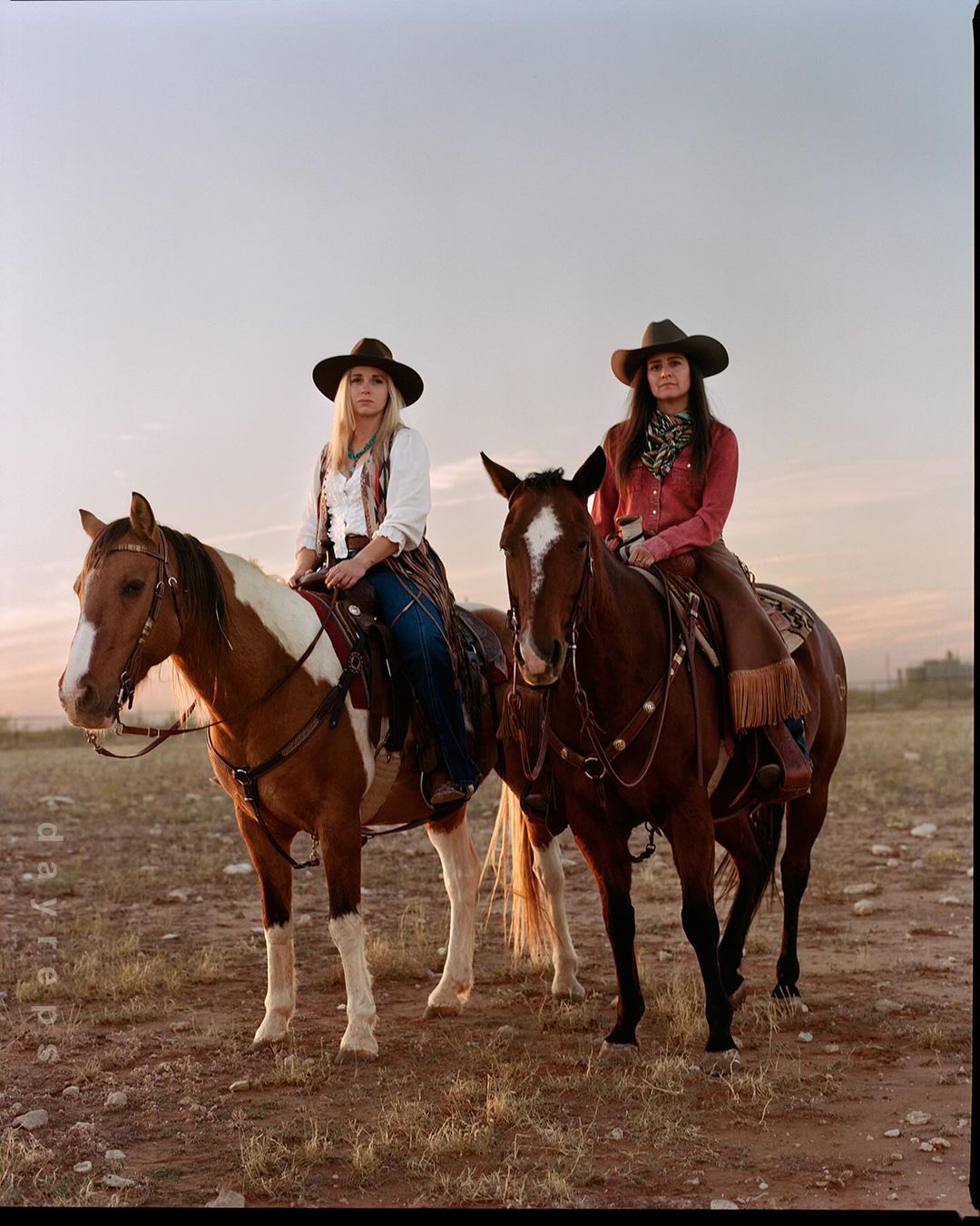 Two women in cowboy hats ride horses in a rural field at sunset. The horses are brown and white, and the women wear Western attire.