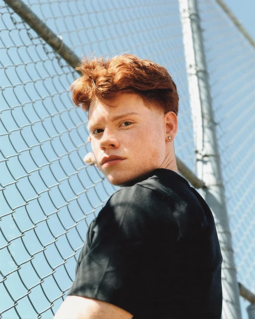 Close-up portrait of a young adult man with red hair looking over his shoulder, wearing a black t-shirt and posing by a chain-link fence.