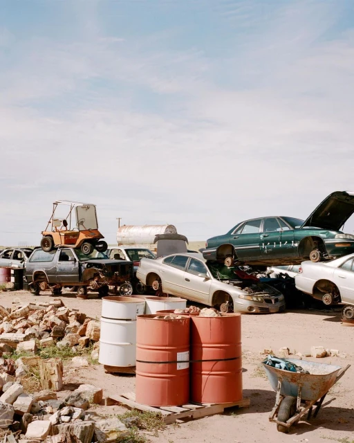 Automobile junkyard filled with wrecked cars, trucks, metal barrels, brick rubble, and a rusted wheelbarrow under a cloudy sky.