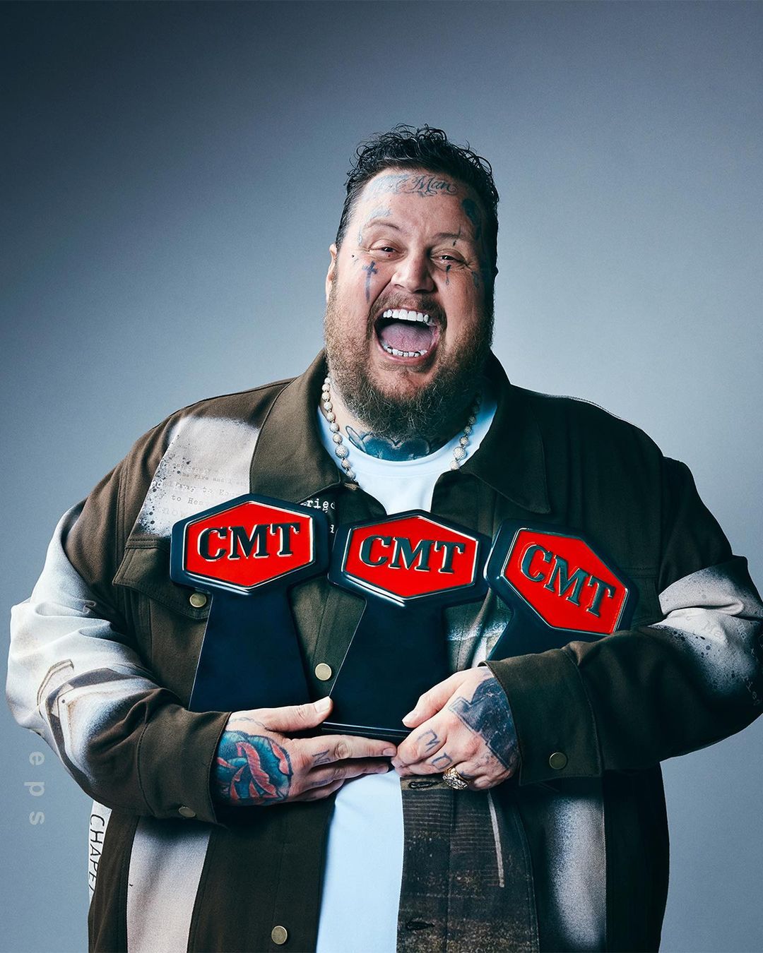 An adult male smiles widely while holding three CMT awards. He has tattoos, a beard, and wears a decorated jacket against a grey background.