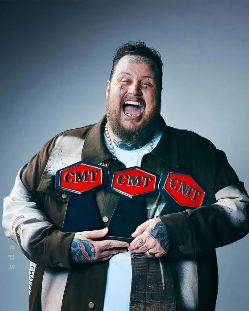 An adult male smiles widely while holding three CMT awards. He has tattoos, a beard, and wears a decorated jacket against a grey background.
