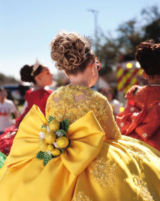 Young girl in a yellow sequined gown with a large bow and decorative lemons, viewed from behind with other girls in the background.