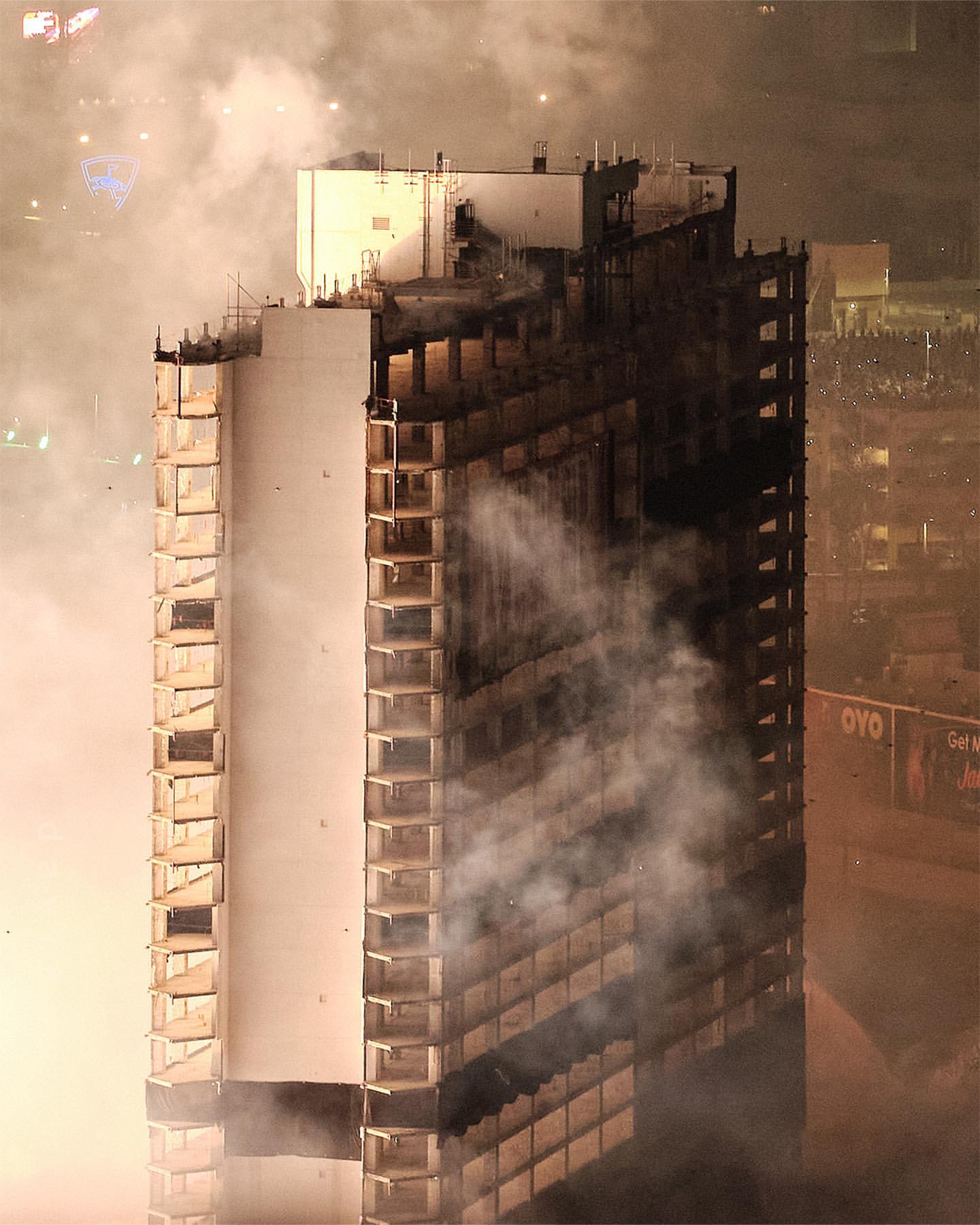 An eye-level shot captures a tall, partially damaged building engulfed in smoke at night, with the night sky barely visible.