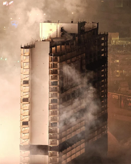 An eye-level shot captures a tall, partially damaged building engulfed in smoke at night, with the night sky barely visible.