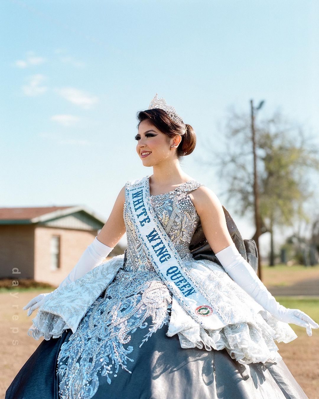 Portrait of a smiling Hispanic woman wearing an ornate gown, tiara, and sash that reads "White Wing Queen" in an outdoor setting.