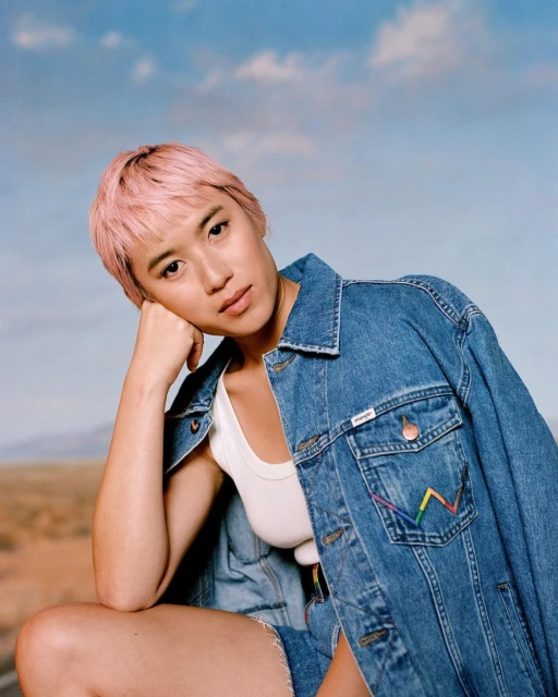 Woman with pink hair wearing a denim jacket with rainbow stitching poses outdoors against a sky and desert background in a medium shot.