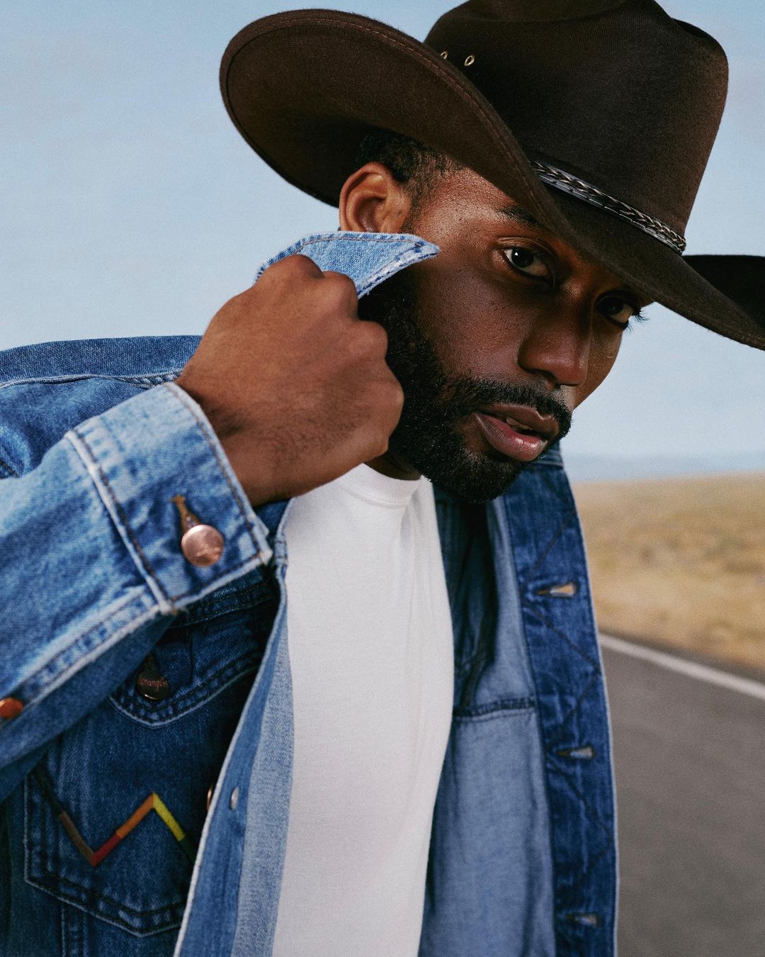 Portrait of an African American man in a cowboy hat and denim jacket standing on a road against a blue sky background.