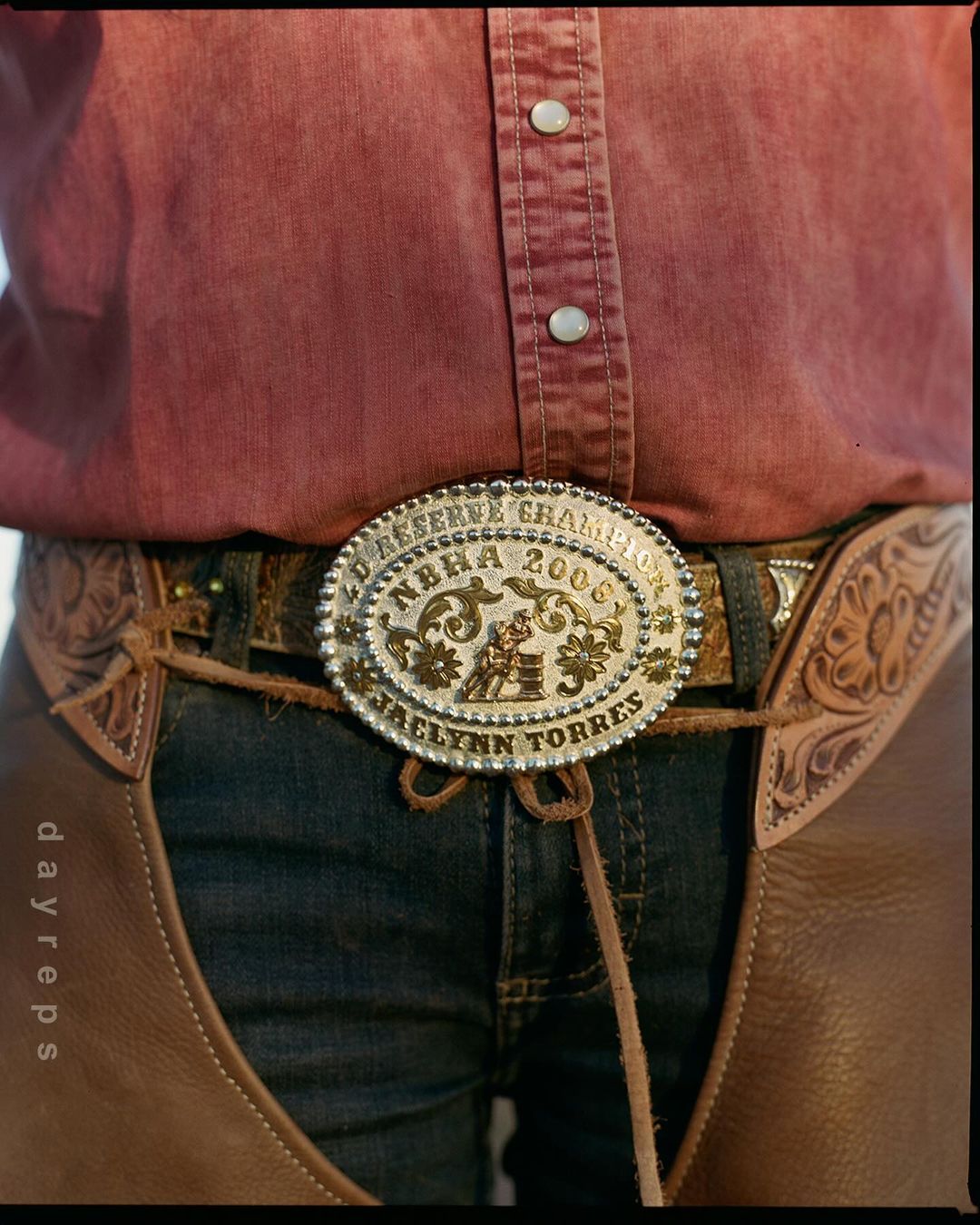 Closeup of a person in a pink shirt, jeans, and leather chaps, wearing a large rodeo belt buckle with the words 'Reserve Champion' on it.