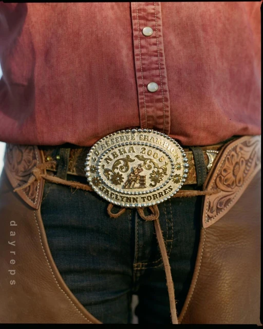 Closeup of a person in a pink shirt, jeans, and leather chaps, wearing a large rodeo belt buckle with the words 'Reserve Champion' on it.