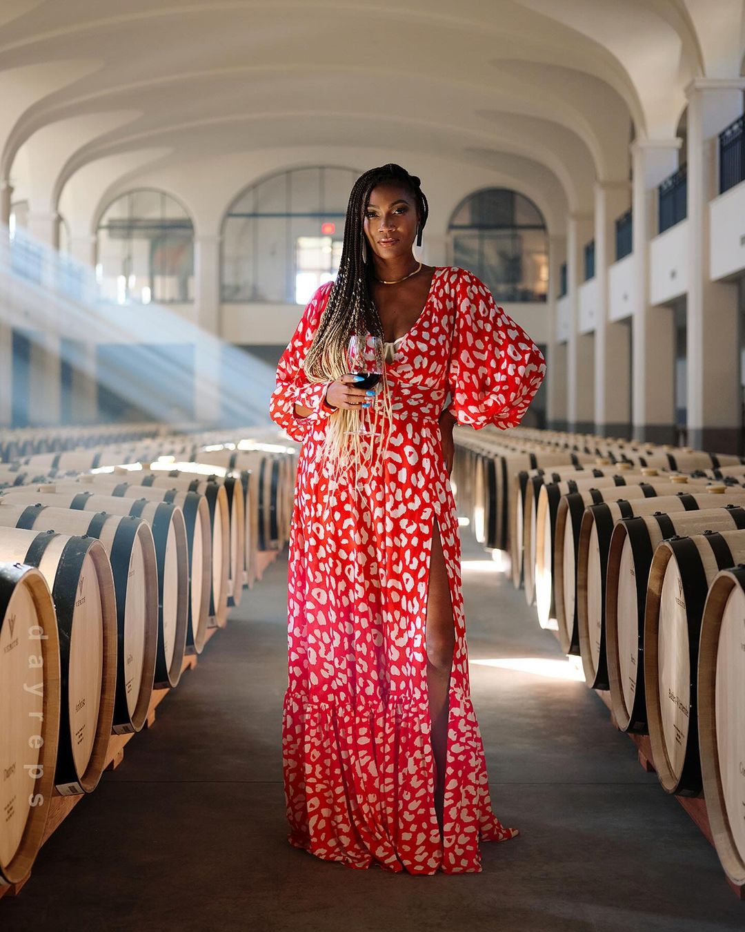An adult woman in a red and white dress poses holding a wine glass in a barrel-filled wine cellar beneath arched ceilings.