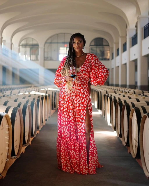 An adult woman in a red and white dress poses holding a wine glass in a barrel-filled wine cellar beneath arched ceilings.