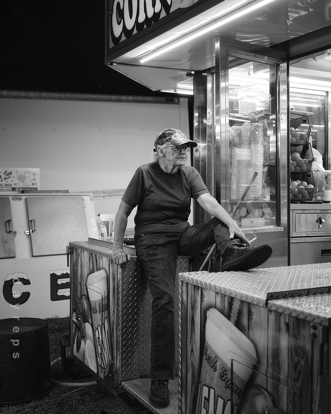 Monochrome shot of an elderly man sitting at a concession stand, wearing a cap and casual clothes, in a fair-like outdoor setting at night.