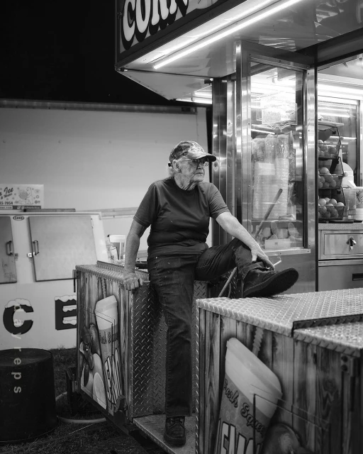Monochrome shot of an elderly man sitting at a concession stand, wearing a cap and casual clothes, in a fair-like outdoor setting at night.